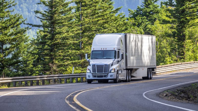 White big rig semi truck transporting goods in refrigerated semi trailer turning on the mountain road with rock wall stock photo