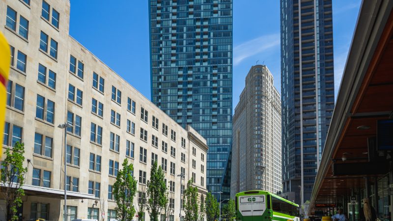 Two tall buildings in the background with a flat iron building in the middle. On the left is a midrise building, and on the right is a green and white GO bus.