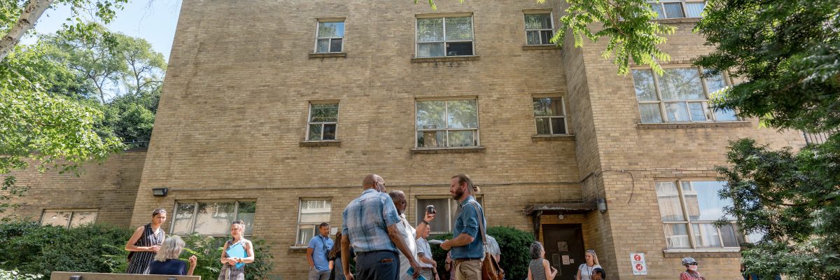 People are gathered in small groups in a green space in front of a multi-level residential building