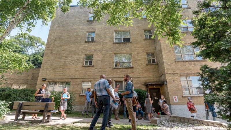 People are gathered in small groups in a green space in front of a multi-level residential building